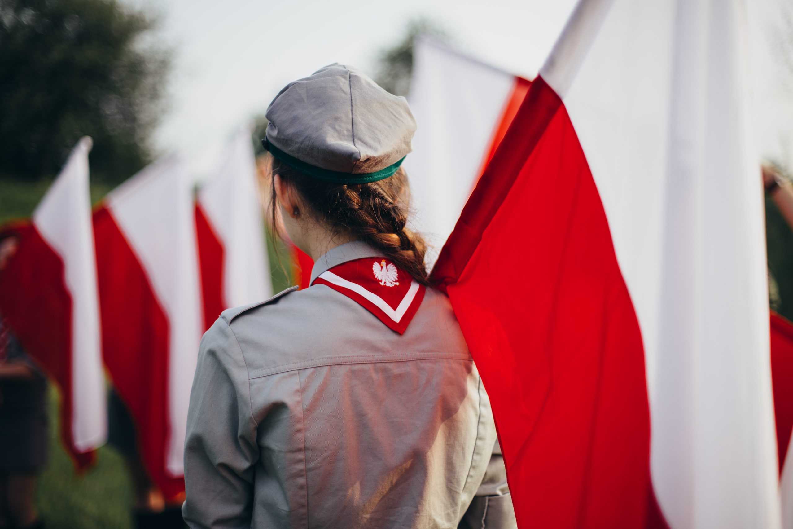 Harcerka trzymająca flagę Polski, odwrócona tyłem;w tle kolejne biało-czerwone flagi. 